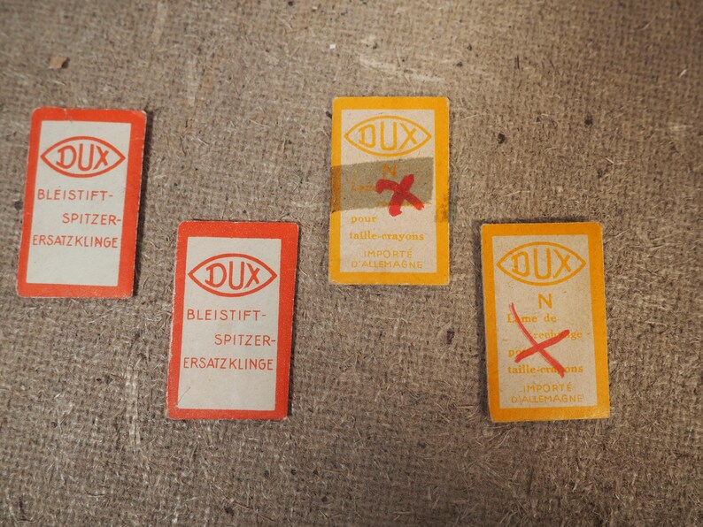 May include: Four vintage DUX pencil sharpener replacement blade cards. Two are red and white, and two are yellow and white. The cards have text in German and French.