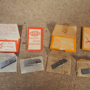 May include: A collection of vintage DUX pencil sharpener replacement blades and packaging. The packaging is orange and white, with German text and illustrations. The blades are silver and rectangular.
