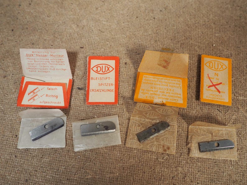 May include: A collection of vintage DUX pencil sharpener replacement blades and packaging. The packaging is orange and white, with German text and illustrations. The blades are silver and rectangular.
