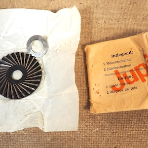 May include: A collection of vintage metal machine parts, including a circular blade with radial grooves and a small washer, alongside a paper package with text in multiple languages. The package is labeled "Jupiter".