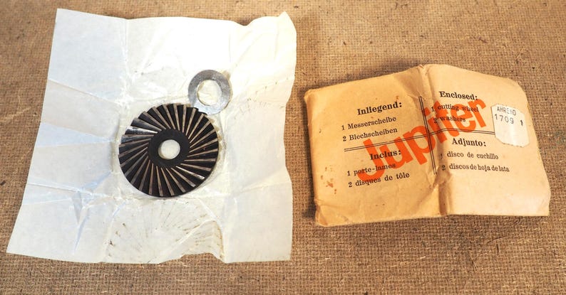 May include: A collection of vintage metal machine parts, including a circular blade with radial grooves and a small washer, alongside a paper package with text in multiple languages. The package is labeled "Jupiter".