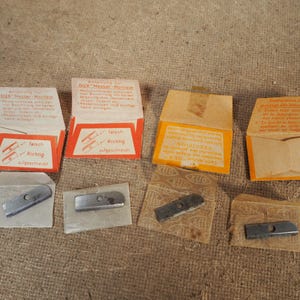 May include: A collection of vintage DUX pencil sharpener blades and packaging. The packaging is orange and white with German text. The blades are silver and rectangular. The packaging includes instructions and diagrams.