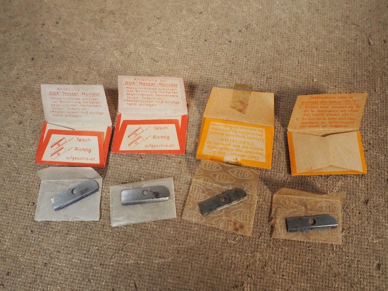 May include: A collection of vintage DUX pencil sharpener blades and packaging. The packaging is orange and white with German text. The blades are silver and rectangular. The packaging includes instructions and diagrams.
