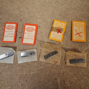 May include: Several DUX pencil sharpener replacement blades and packaging are displayed. The packaging is rectangular with the DUX logo and text in German. The blades are silver and rectangular. Some packaging has a red "X" mark.