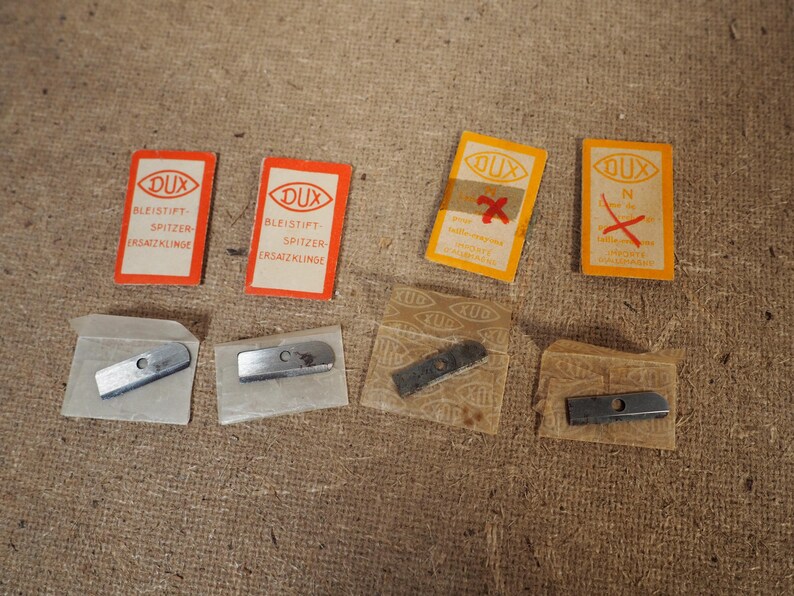 May include: Several DUX pencil sharpener replacement blades and packaging are displayed. The packaging is rectangular with the DUX logo and text in German. The blades are silver and rectangular. Some packaging has a red "X" mark.