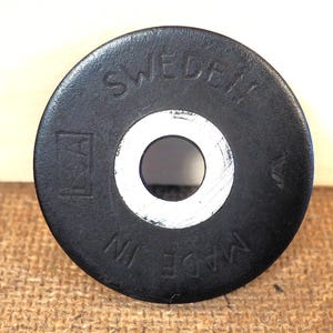 May include: A black, circular object with a central hole and the text "SWEDEN" and "MADE IN" in white lettering. The object has a white inner ring and a logo on the left side.