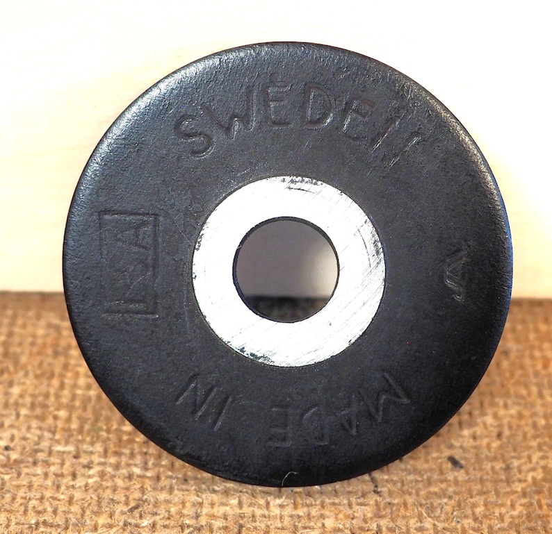 May include: A black, circular object with a central hole and the text "SWEDEN" and "MADE IN" in white lettering. The object has a white inner ring and a logo on the left side.