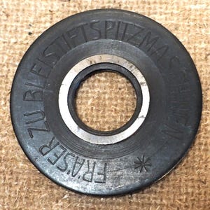 May include: A circular, dark gray metal part with a central hole and a silver ring. The outer edge features embossed text, including the word "FRASER". A small asterisk is also visible. The part appears to be a component of a machine.