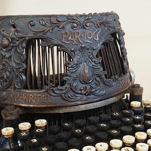 May include: Vintage black typewriter with an elaborate metal cover, engraved with "BAR-LOCK" and "TYPEWRITER". The keys are black and cream. A clear glass bottle with a metal clasp is in the background.