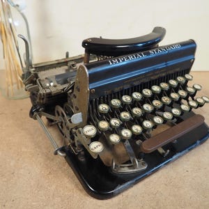 May include: A black Imperial Standard typewriter with a brown carriage return lever. The typewriter has a standard QWERTY keyboard with keys labelled "Caps" and "Figs".