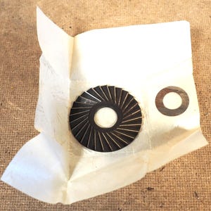 May include: A dark brown, circular metal component with radial grooves, and a smaller, flat, circular metal washer. Both parts are on a piece of crumpled white paper, set against a textured, brown background.