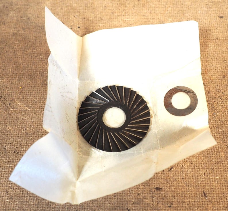 May include: A dark brown, circular metal component with radial grooves, and a smaller, flat, circular metal washer. Both parts are on a piece of crumpled white paper, set against a textured, brown background.