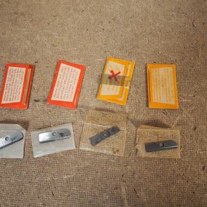 May include: An assortment of vintage metal pencil sharpener blades and packaging. The packaging includes orange and yellow rectangular boxes with text, and the blades are in small, clear plastic sleeves.