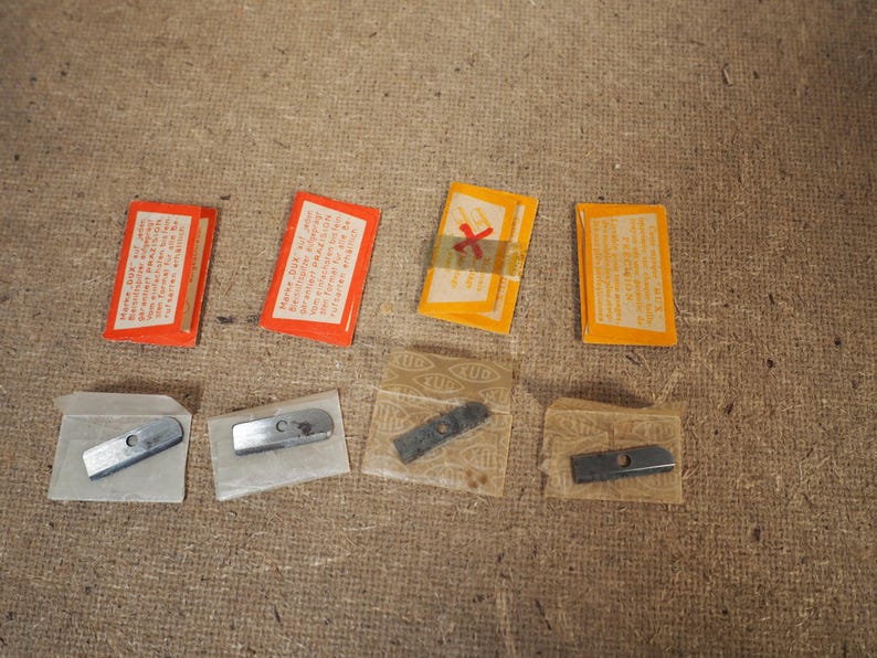 May include: An assortment of vintage metal pencil sharpener blades and packaging. The packaging includes orange and yellow rectangular boxes with text, and the blades are in small, clear plastic sleeves.