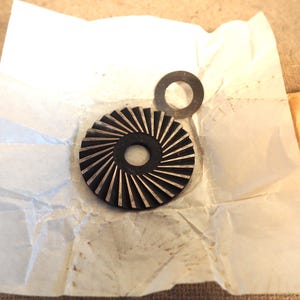 May include: A black and silver gear-shaped component with radial grooves, and a small, circular washer. The parts rest on a crumpled white paper, with a tan paper visible in the background. The gear has a central hole.