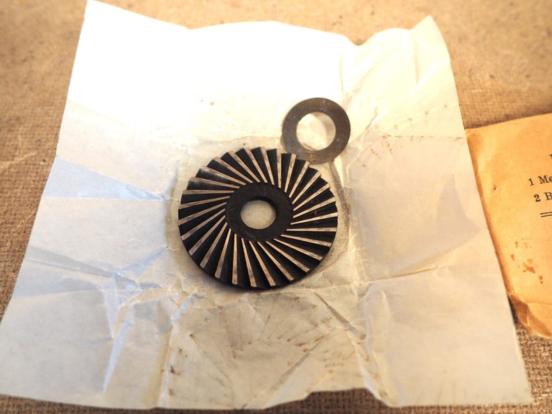 May include: A black and silver gear-shaped component with radial grooves, and a small, circular washer. The parts rest on a crumpled white paper, with a tan paper visible in the background. The gear has a central hole.