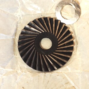 May include: A close-up of two metal machine parts. One is a dark, circular gear with angled teeth and a central hole. The other is a silver, circular washer. Both parts are on a crumpled, off-white paper surface.