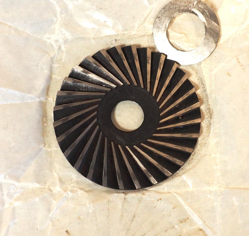 May include: A close-up of two metal machine parts. One is a dark, circular gear with angled teeth and a central hole. The other is a silver, circular washer. Both parts are on a crumpled, off-white paper surface.