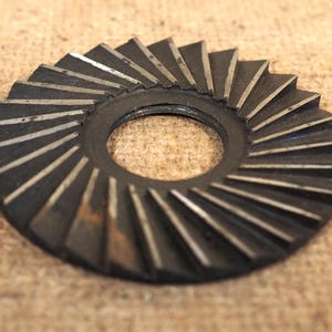 May include: A circular, dark metal gear with angled teeth and a central hole. The gear's teeth have a silver-colored edge, and the overall design suggests a mechanical or industrial component.