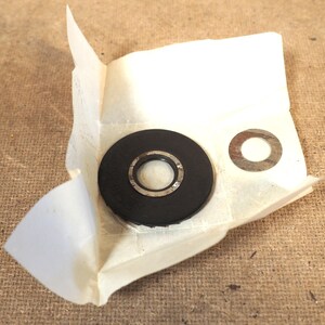 May include: A black rubber washer with a silver metal ring is partially unwrapped from a piece of paper. A separate silver metal washer is also visible. The items are on a brown surface.