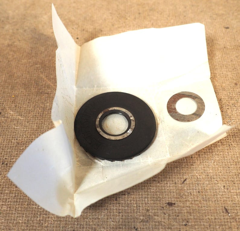 May include: A black rubber washer with a silver metal ring is partially unwrapped from a piece of paper. A separate silver metal washer is also visible. The items are on a brown surface.