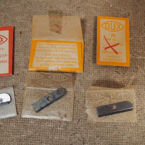 May include: A collection of vintage DUX pencil sharpener replacement blades and packaging. The packaging includes orange and red rectangular cards with text in German and French. The blades are silver and rectangular.