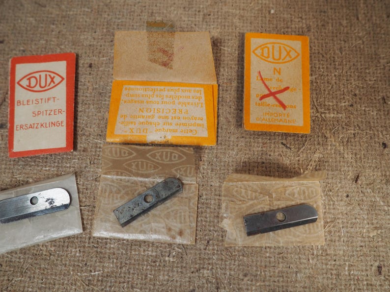 May include: A collection of vintage DUX pencil sharpener replacement blades and packaging. The packaging includes orange and red rectangular cards with text in German and French. The blades are silver and rectangular.