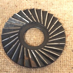 May include: A circular, dark metal washer with a central hole and a series of angled, silver-colored blades radiating outwards. The blades are evenly spaced, creating a fan-like pattern. The washer appears to be used for industrial purposes.