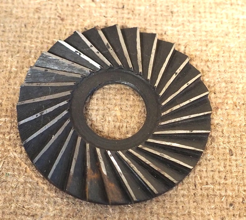 May include: A circular, dark metal washer with a central hole and a series of angled, silver-colored blades radiating outwards. The blades are evenly spaced, creating a fan-like pattern. The washer appears to be used for industrial purposes.