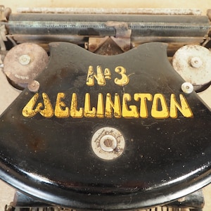 May include: Close-up of a vintage black typewriter featuring the text "No 3 WELLINGTON" in yellow. The typewriter has a curved design with metal parts and circular spools on either side.