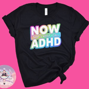May include: Black t-shirt featuring the phrase "NOW THAT'S WHAT I CALL ADHD" in a vibrant, outlined font. The text is stacked, with the words arranged vertically. The shirt has rolled sleeves and a round neckline.