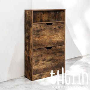 K&ouml;nnte beinhalten: Ein brauner Schuhschrank aus Holz mit zwei Schubladen und einem Regal. Der Schuhschrank hat eine Antik-Holz-Optik und steht auf einem wei&szlig;en Fliesenboden.