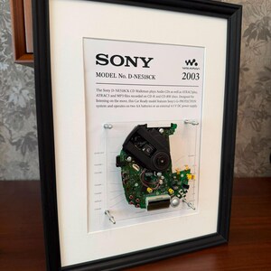 Pode incluir: Uma moldura exibindo a placa de circuito interna de um Sony D-NE518CK Walkman. A placa está montada num painel de acrílico transparente dentro de uma moldura preta, acompanhada de texto impresso detalhando as especificações do modelo de 2003.