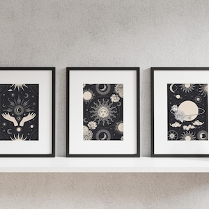 Peut inclure: Trois impressions encadrées noires avec des motifs célestes. Les impressions présentent un soleil, une lune, des étoiles et des mains tenant un orbe céleste. Les impressions sont sur une étagère blanche contre un mur gris.
