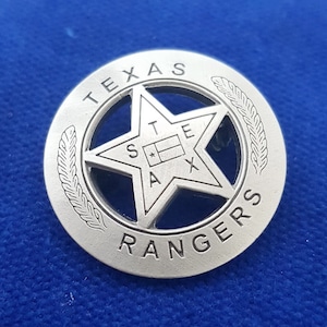 Könnte beinhalten: Eine silberfarbene Metallnadel mit einem fünfzackigen Stern in der Mitte. Der Stern trägt die Inschriften "STATE" und "TEXAS" im Inneren. Die Nadel ist von einem Kreis umgeben, auf dem die Worte "TEXAS RANGERS" eingraviert sind.