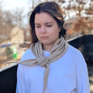 May include: A beige knitted scarf tied in a knot, with long ends hanging down. The scarf is worn over a white long-sleeved shirt. The background is blurred, showing trees and a building.