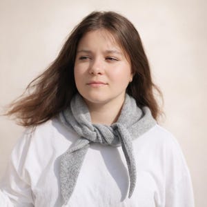 May include: A light grey knitted scarf is draped around the neck. The scarf is made of a soft, textured material and is tied in a knot at the front. The person is wearing a white shirt.