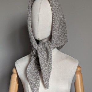 Puede incluir: Un conjunto de capucha y bufanda de punto gris exhibido en un maniqu&iacute;. La capucha est&aacute; dise&ntilde;ada para cubrir la cabeza y el cuello, con la bufanda anudada en la parte delantera. La prenda de punto tiene una apariencia texturizada.