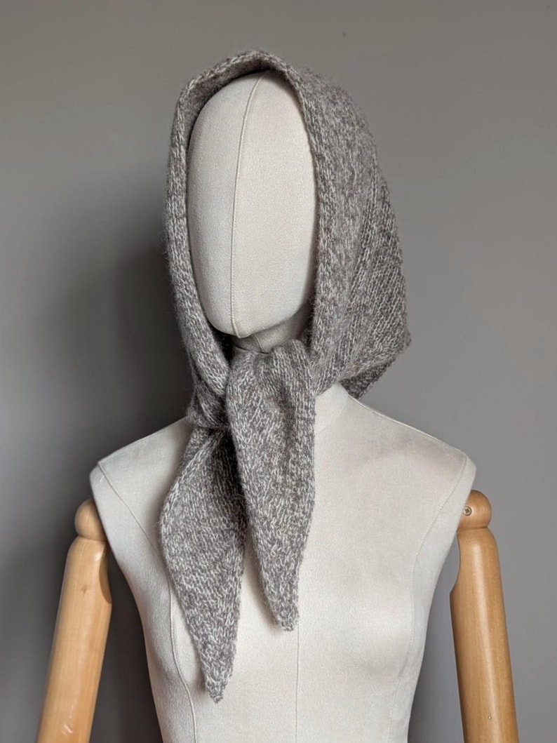 Puede incluir: Un conjunto de capucha y bufanda de punto gris exhibido en un maniqu&iacute;. La capucha est&aacute; dise&ntilde;ada para cubrir la cabeza y el cuello, con la bufanda anudada en la parte delantera. La prenda de punto tiene una apariencia texturizada.