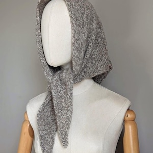 Puede incluir: Conjunto de capucha y bufanda de punto gris, exhibido en un maniqu&iacute;. La capucha est&aacute; dise&ntilde;ada para cubrir la cabeza y el cuello, con la bufanda atada en la parte delantera. El tejido de punto tiene un aspecto texturizado, lo que sugiere un material c&aacute;lido y acogedor.