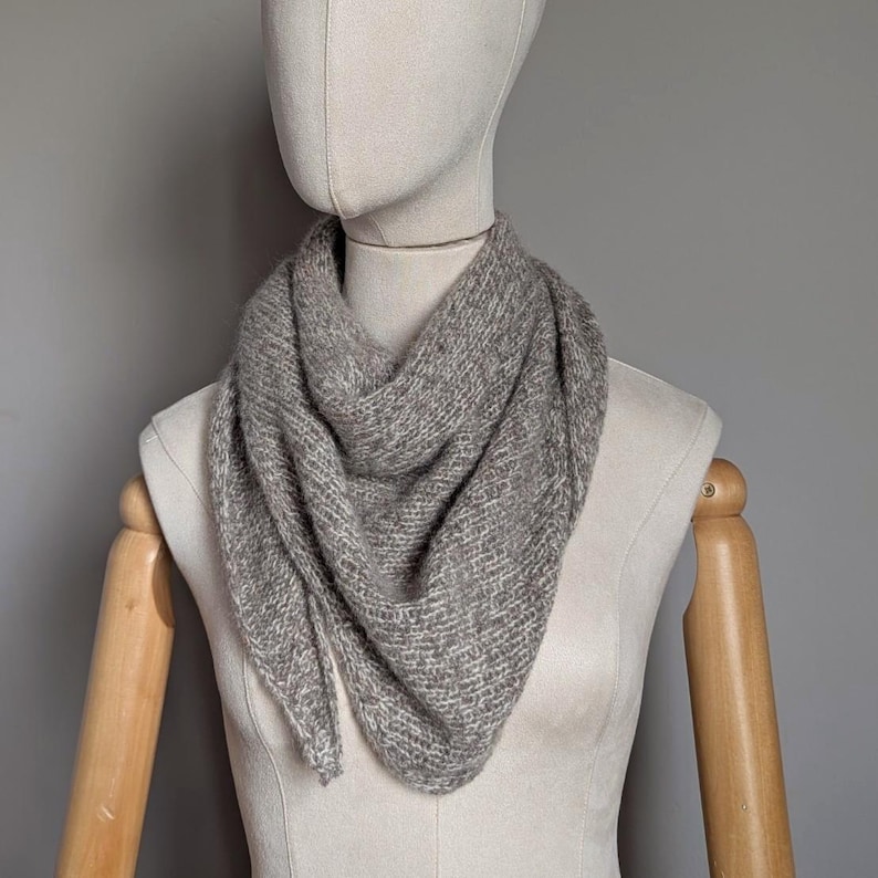 Puede incluir: Una bufanda triangular gris claro hecha de tejido de punto suave y texturizado. La bufanda est&aacute; drapeada alrededor del cuello de un maniqu&iacute;, mostrando su forma y ca&iacute;da. El color neutro lo convierte en un accesorio vers&aacute;til.