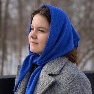 May include: A woman wearing a vibrant blue knitted headscarf and a gray wool coat. The headscarf is draped over the head and tied around the neck. The coat has a classic design with large lapels. The background is blurred, suggesting an outdoor setting.