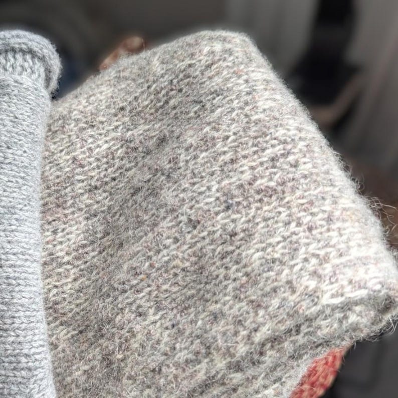 Puede incluir: Primer plano de una tela de punto gris claro doblada. La prenda de punto tiene un aspecto texturizado con motas de fibras m&aacute;s oscuras y claras. El borde de una manga enrollada es visible en el lado izquierdo de la imagen.