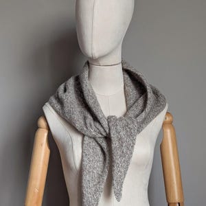 Puede incluir: Una bufanda gris tejida est&aacute; drapeada alrededor del cuello de un maniqu&iacute;. La bufanda est&aacute; atada con un nudo en la parte delantera, con los extremos colgando. La bufanda tiene un aspecto texturizado y est&aacute; hecha de un material suave y c&aacute;lido.