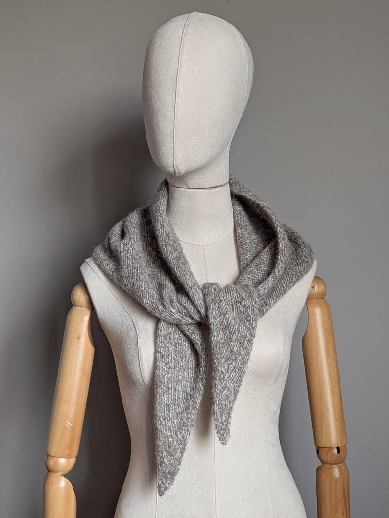 Puede incluir: Una bufanda gris tejida est&aacute; drapeada alrededor del cuello de un maniqu&iacute;. La bufanda est&aacute; atada con un nudo en la parte delantera, con los extremos colgando. La bufanda tiene un aspecto texturizado y est&aacute; hecha de un material suave y c&aacute;lido.