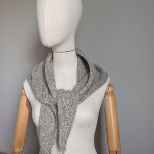 Puede incluir: Una bufanda de punto gris triangular est&aacute; drapeada alrededor del cuello de un maniqu&iacute;. La bufanda tiene una apariencia texturizada y un extremo puntiagudo. El maniqu&iacute; tiene una cara neutra y brazos de madera. El fondo es gris liso.
