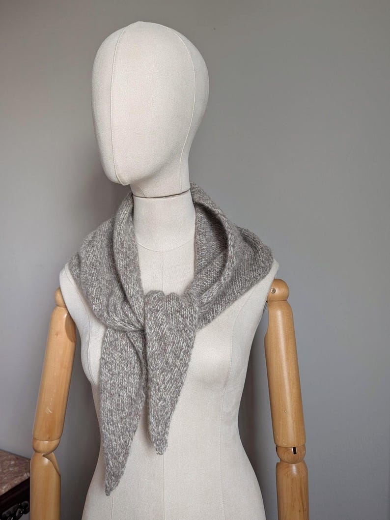 Puede incluir: Una bufanda de punto gris triangular est&aacute; drapeada alrededor del cuello de un maniqu&iacute;. La bufanda tiene una apariencia texturizada y un extremo puntiagudo. El maniqu&iacute; tiene una cara neutra y brazos de madera. El fondo es gris liso.