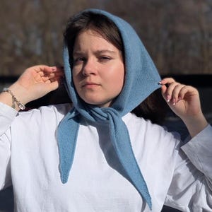 May include: A light blue knitted headscarf, tied under the chin. The scarf is made of a soft, textured material and is worn over the head. The scarf is a solid light blue color and is worn with a white shirt.