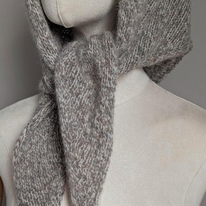 Puede incluir: Una bufanda gris tejida con capucha, exhibida en un maniqu&iacute;. La bufanda est&aacute; hecha de un hilo suave y texturizado con una mezcla de fibras grises y blancas. La bufanda est&aacute; atada al cuello, con la capucha sobre la cabeza.