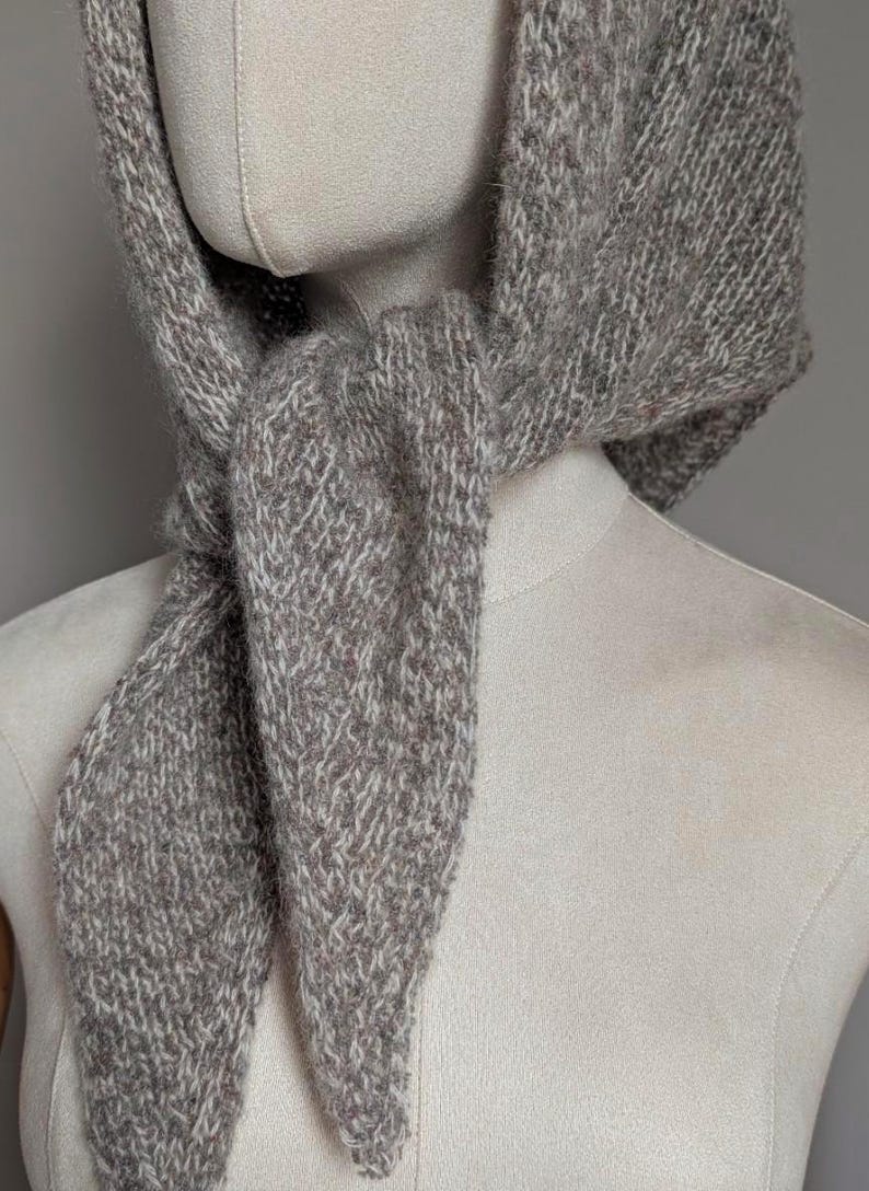 Puede incluir: Una bufanda gris tejida con capucha, exhibida en un maniqu&iacute;. La bufanda est&aacute; hecha de un hilo suave y texturizado con una mezcla de fibras grises y blancas. La bufanda est&aacute; atada al cuello, con la capucha sobre la cabeza.